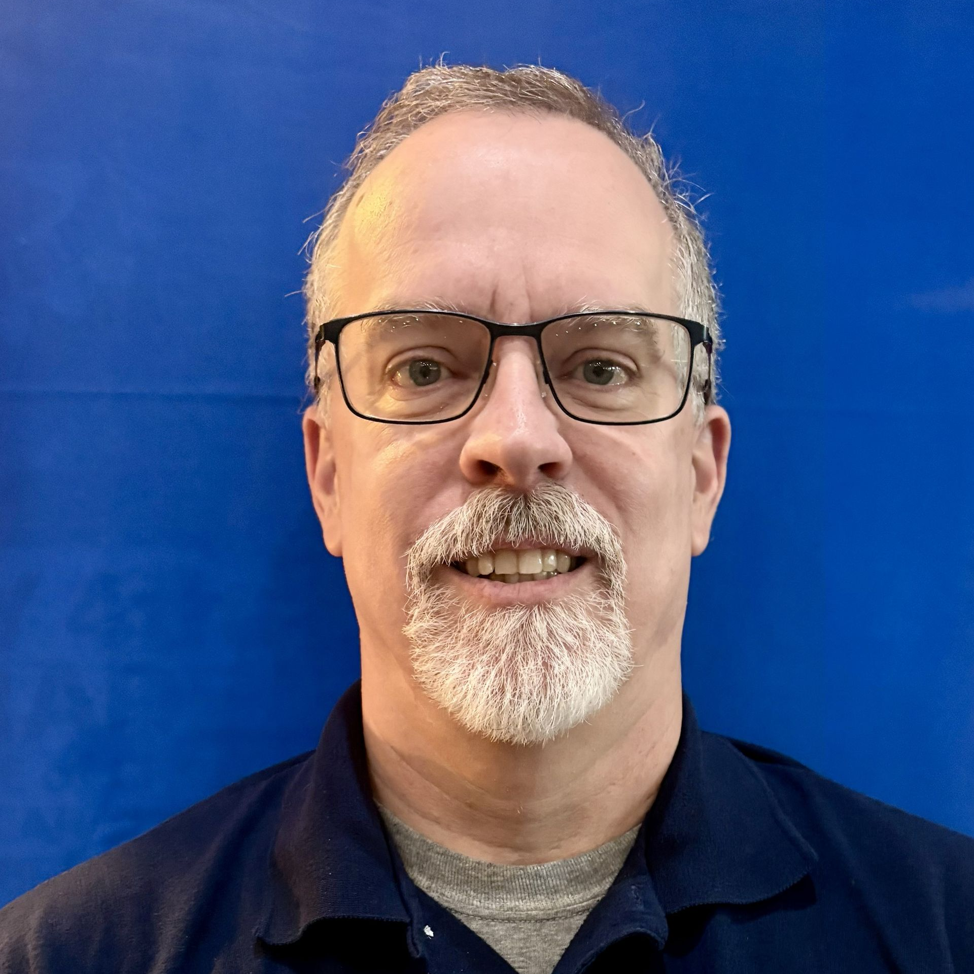 Man with glasses and grey beard smiling against a blue background.