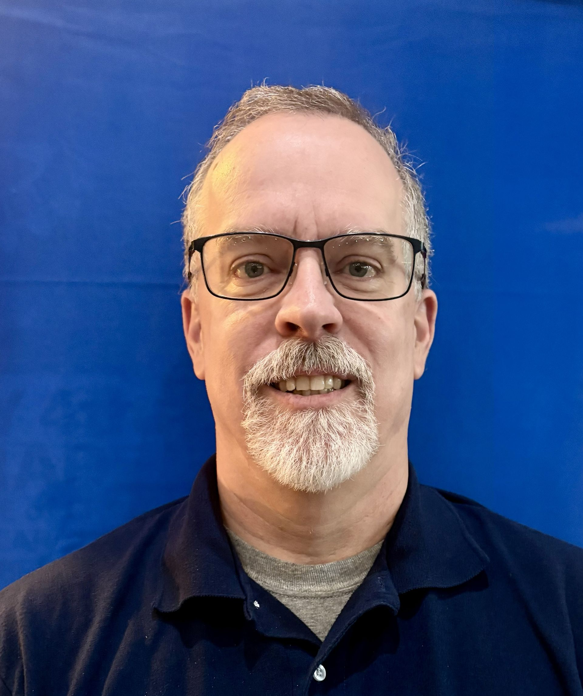 Man with glasses and grey beard smiling, wearing a dark blue shirt against a blue backdrop.