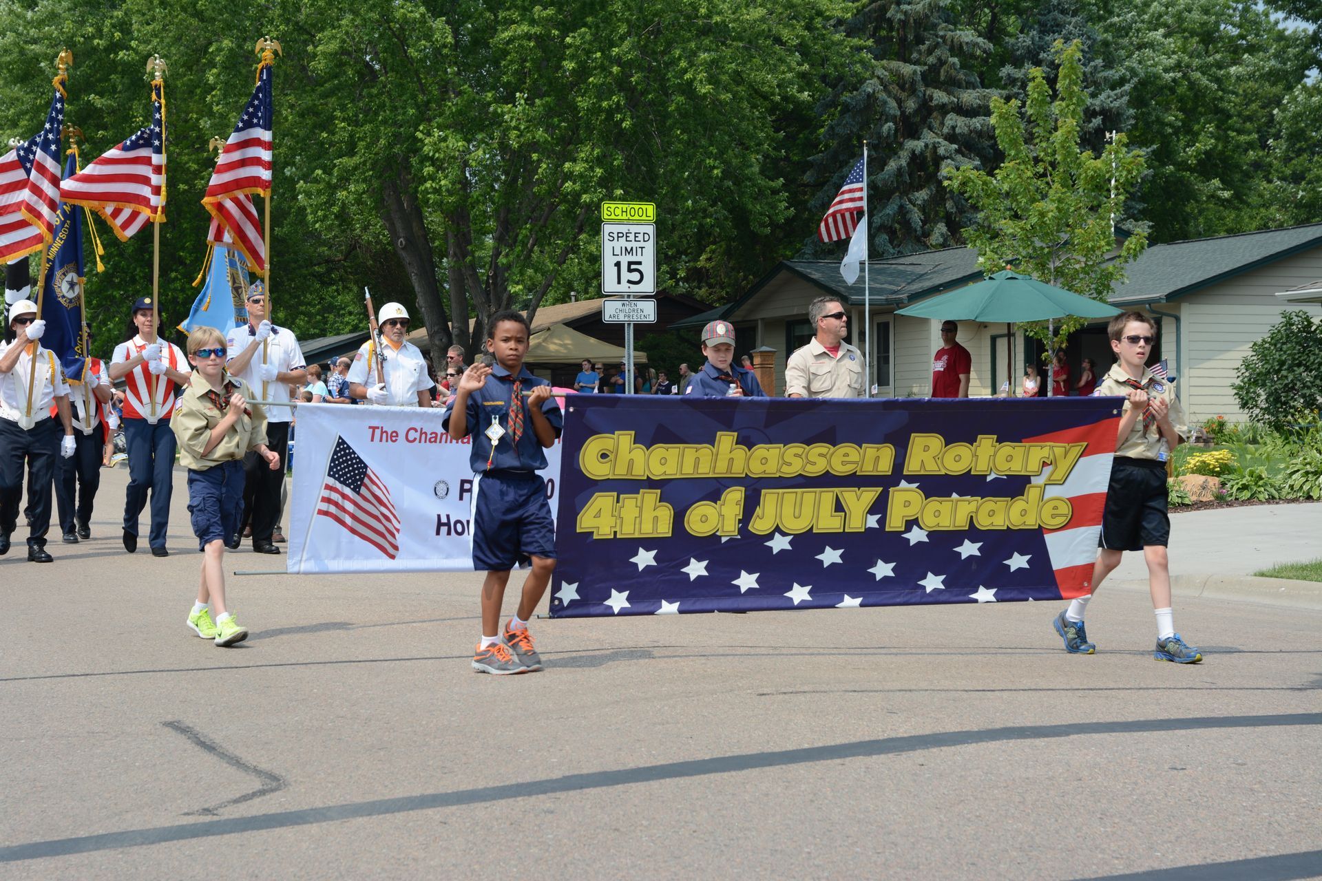 Chanhassen Rotary 4th of July parade with banner, flag bearers, and boy scouts marching on street.