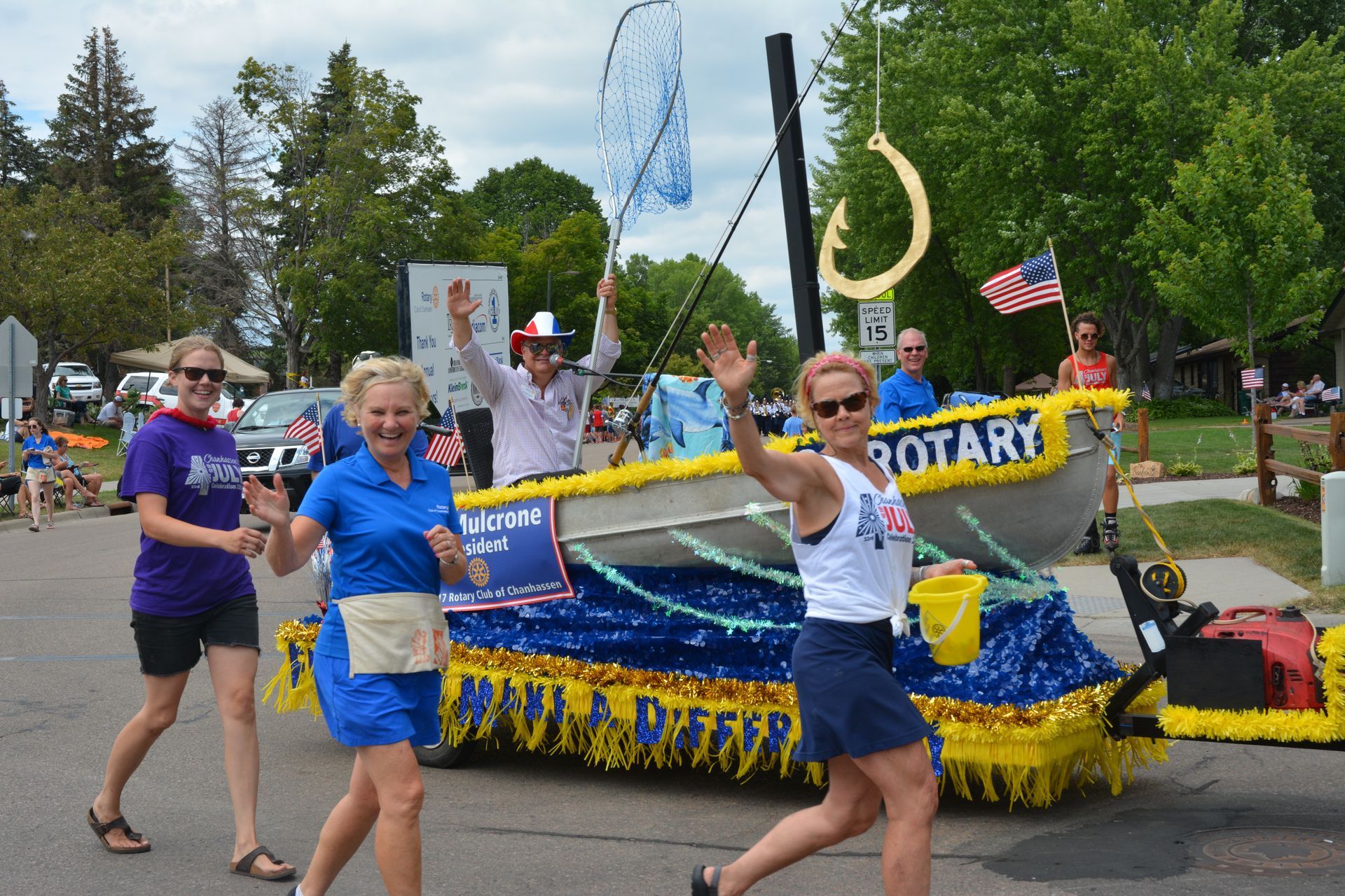 People wave from a decorated boat-shaped float with a fishing hook. Parade setting, sunny day.