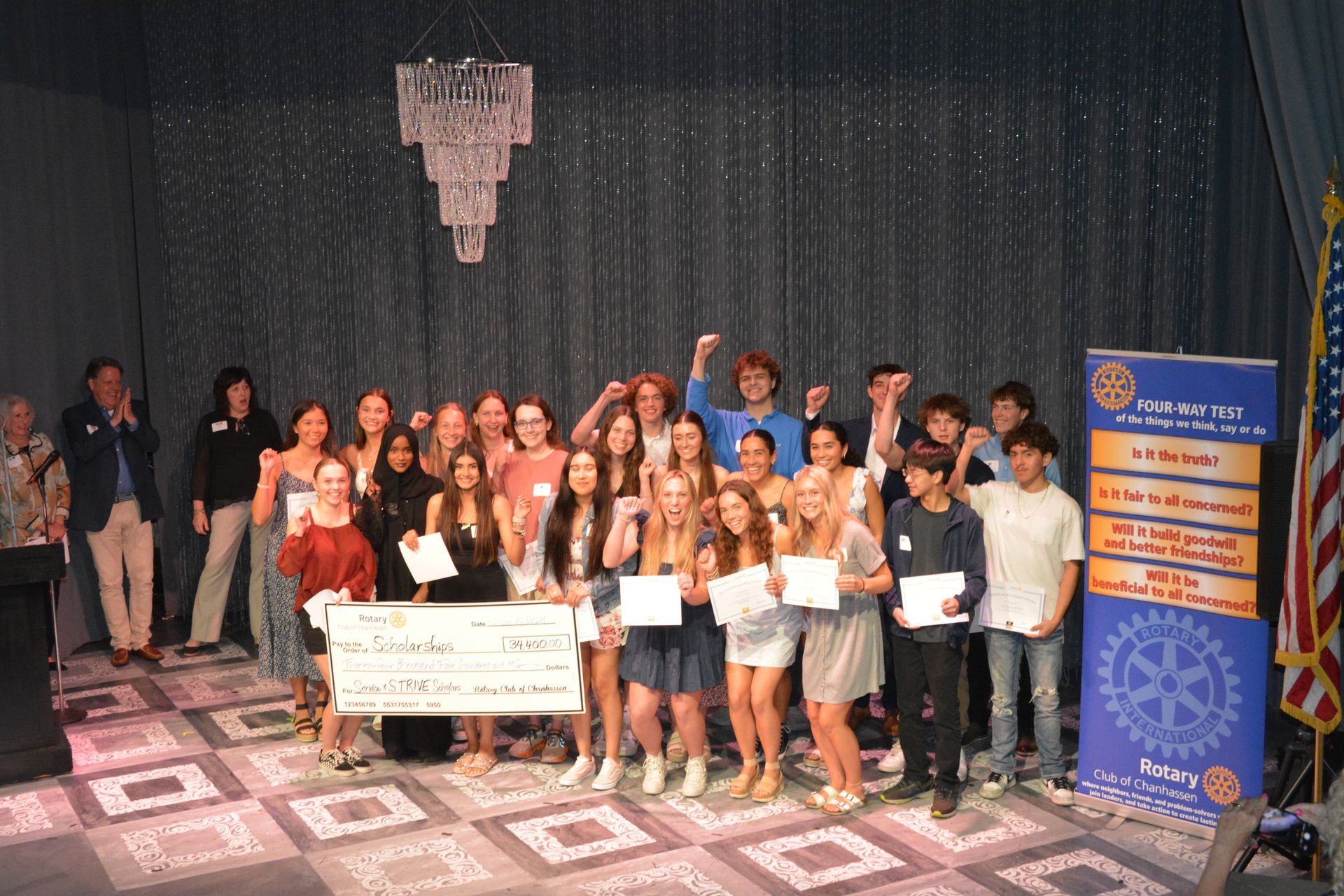 Group of people holding certificates and a large check in a room with a stage, celebratory.