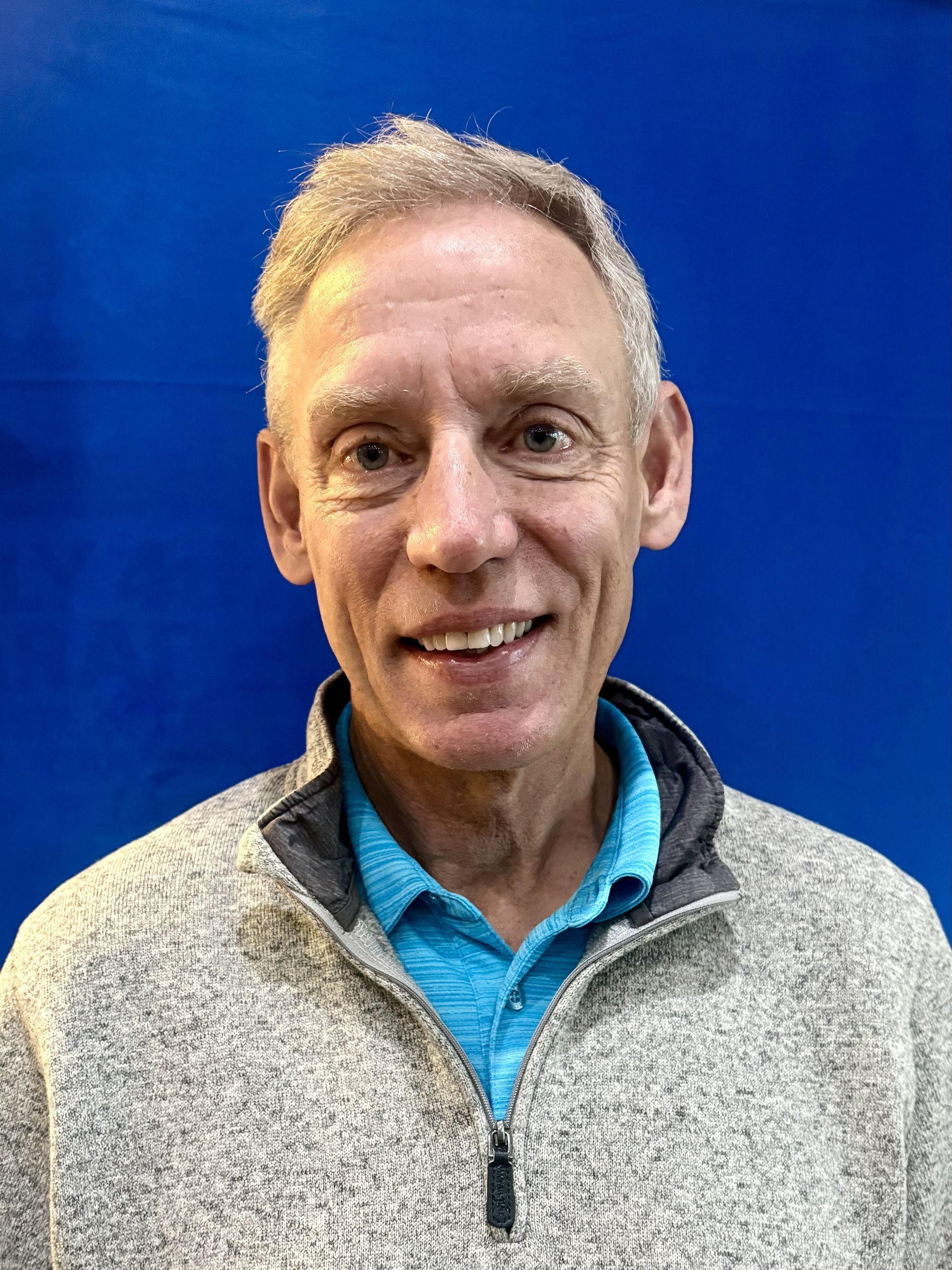 Man smiles, wearing grey sweater over a teal shirt, blue background.