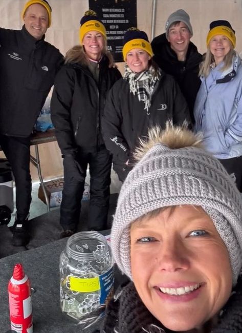 Group of six people in winter hats and jackets smiling indoors. One is taking a selfie.
