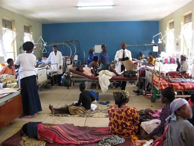 Hospital room with patients in beds, staff attending. People seated on floor and beds. Blue walls.