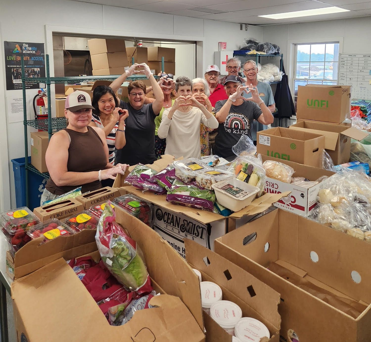People volunteer at a food bank, forming hearts with their hands, surrounded by food boxes.