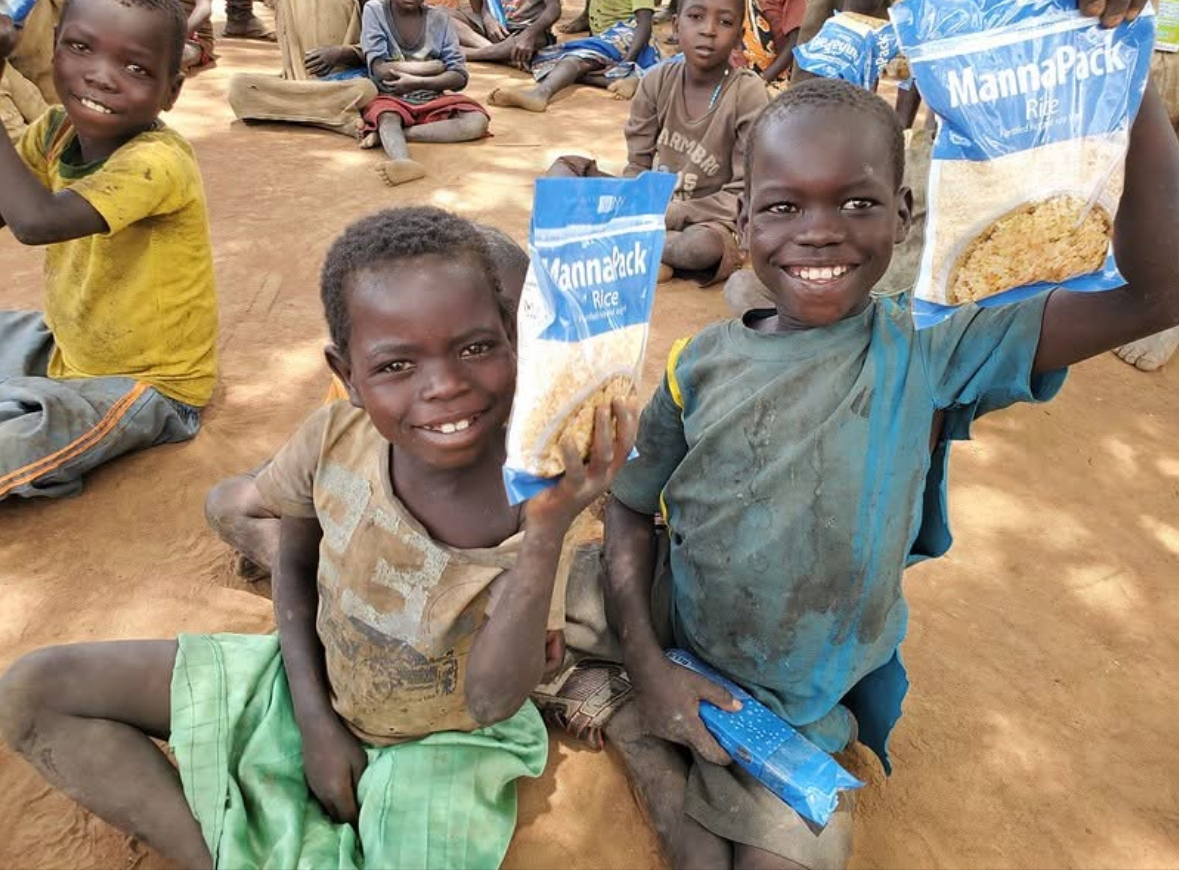 Two children smiling, holding bags of food, sitting outdoors with other children.