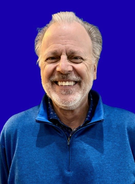 Smiling man with gray hair and beard, wearing a blue zip-up sweater, against a blue background.