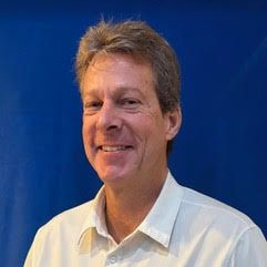 Man smiling, wearing a light-colored button-down shirt, against a blue background.