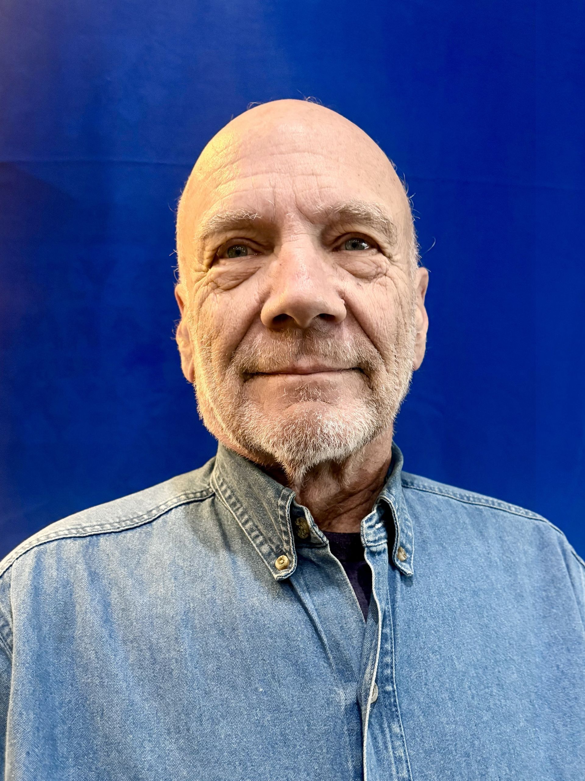 Bald man wearing a denim shirt, smiling, in front of a blue background.