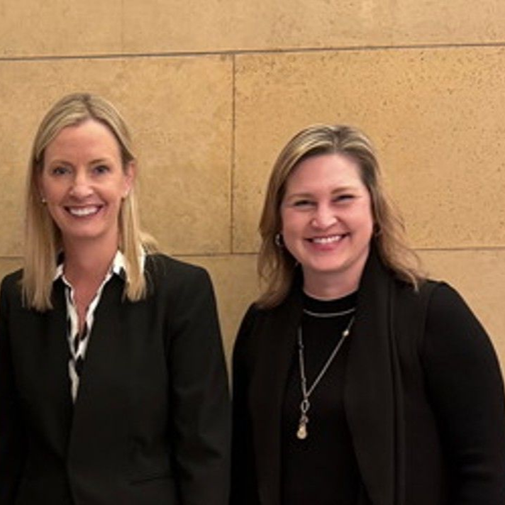 Two smiling women in business attire pose in front of a tan wall.