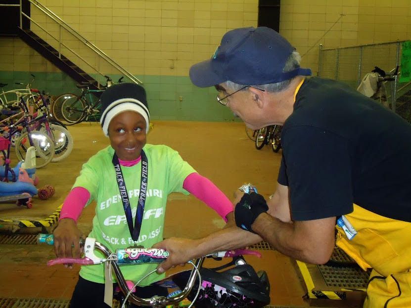 Girl smiles, gripping bike handles while man adjusts them. Indoors, bikes in background. Girl wears green shirt, hat, and pink sleeves.