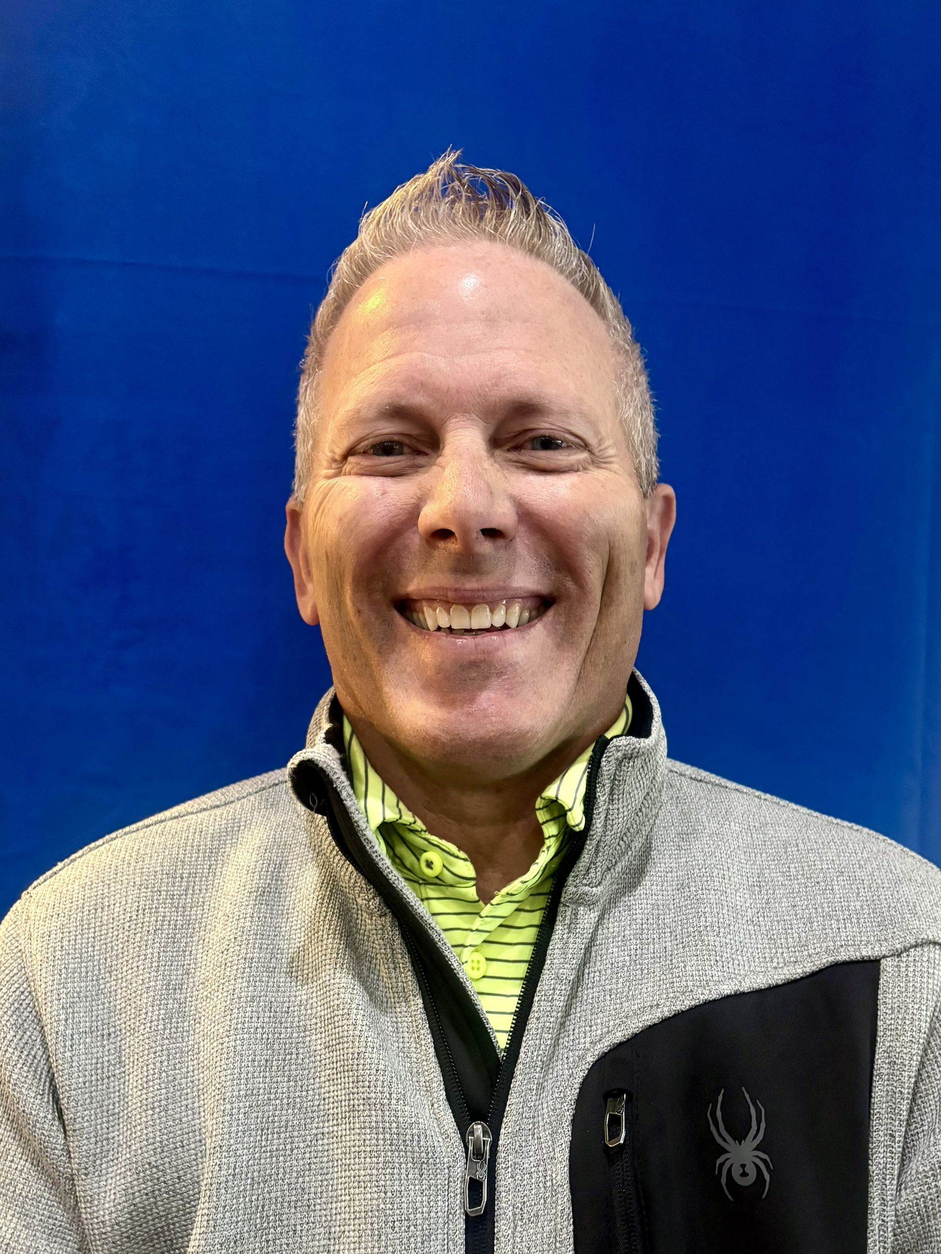 Man with light hair smiles wearing a grey and black jacket over a green shirt, blue background.
