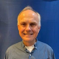 Smiling man with light skin wearing a blue checkered shirt, against a blue background.