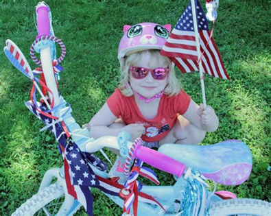 Child wearing a pink cat helmet, sunglasses, and red shirt, holding an American flag. Decorated bike.