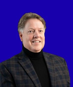 Man in black turtleneck and plaid blazer smiling against a blue background.