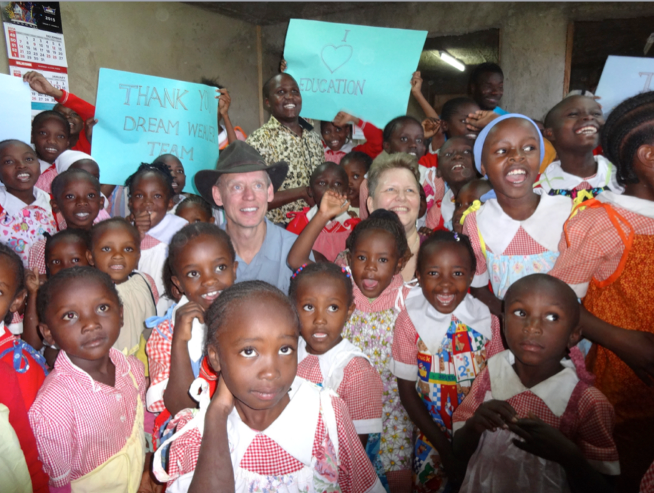 Group of children with two adults holding signs; indoors. Many are smiling, wearing school uniforms.
