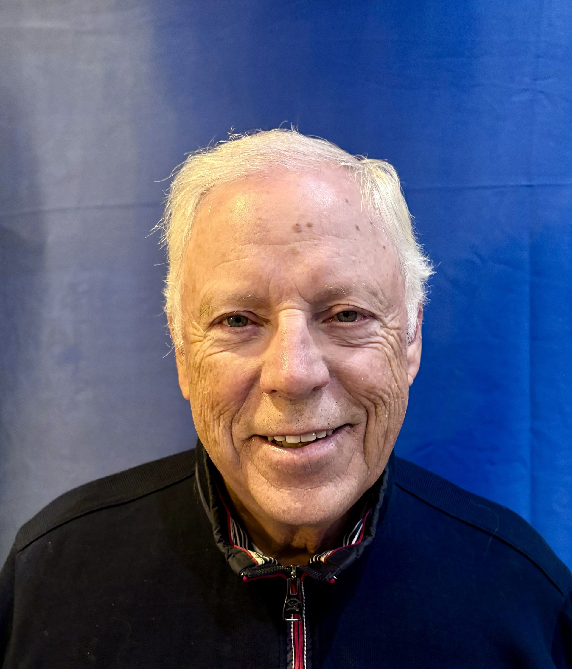 Man with short white hair, smiling in front of a blue background, wearing a black jacket.
