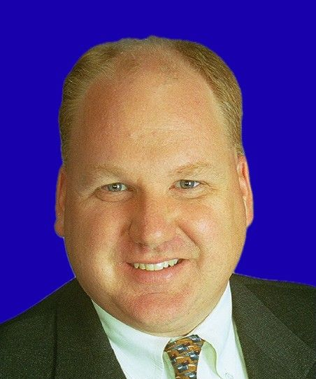 Man with light skin, smiling, in a suit, against a blue background.