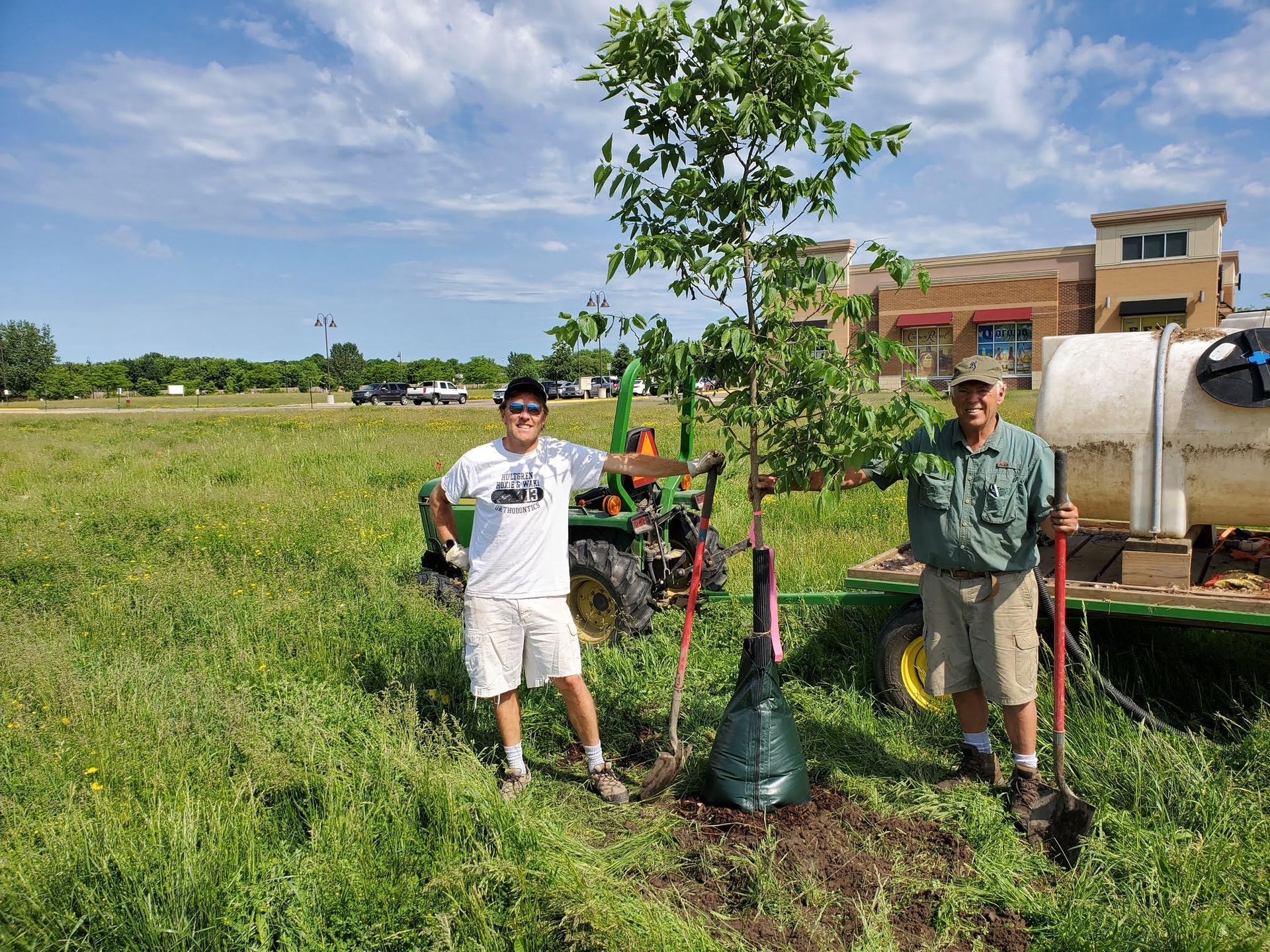 Two people stand by a newly planted tree in a field; one holds a shovel, a tractor is nearby.