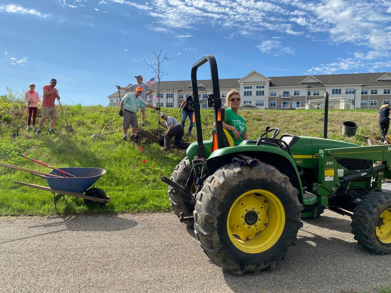 People work on a grassy hillside, a tractor in the foreground, blue sky.