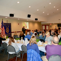 Rotary Club meeting; people seated at tables, speaker at podium showing a slide, American flag in background.