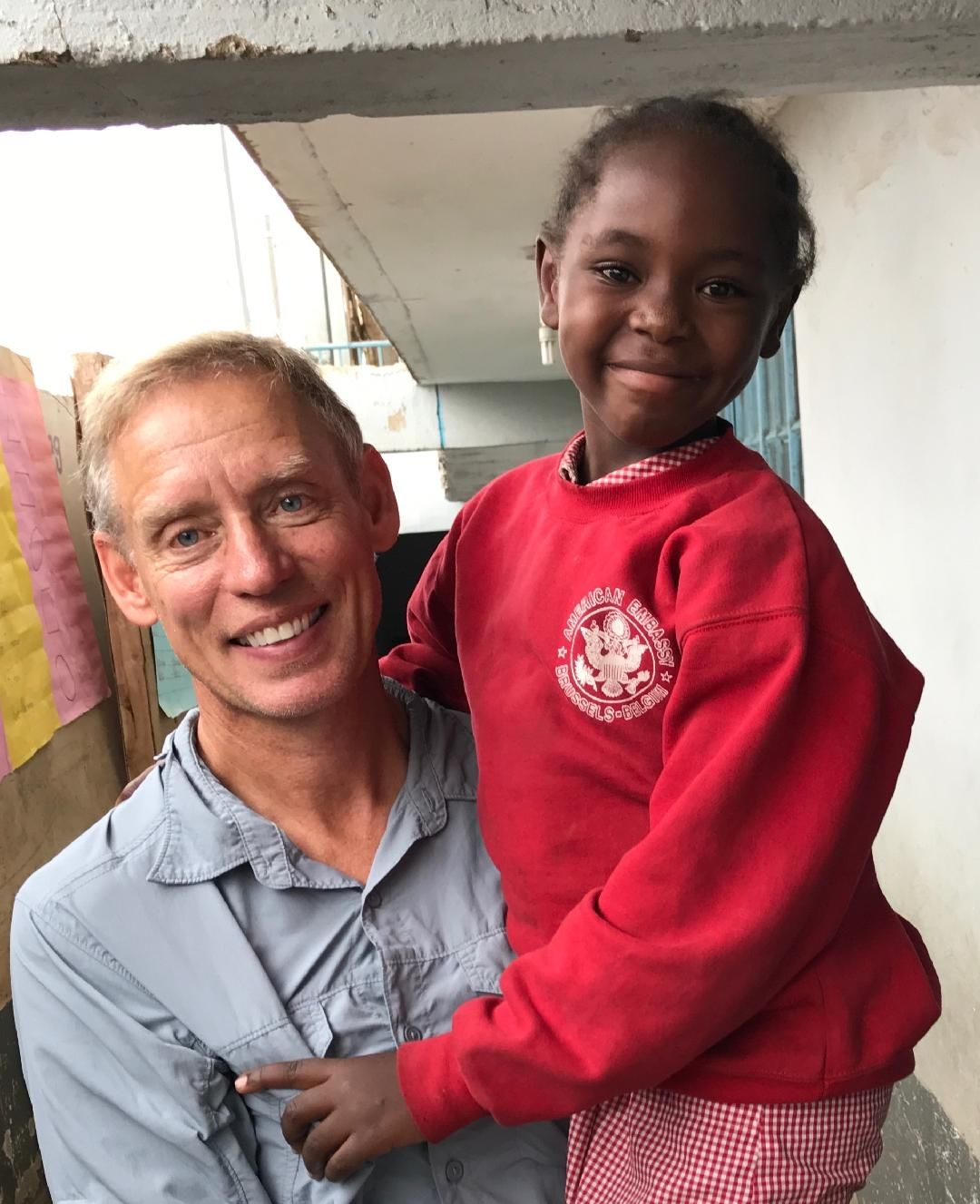Man smiles, holding a girl in red sweater. They pose together outside a building.
