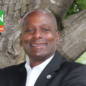 Smiling man in black suit jacket and white shirt standing outdoors by a tree trunk.