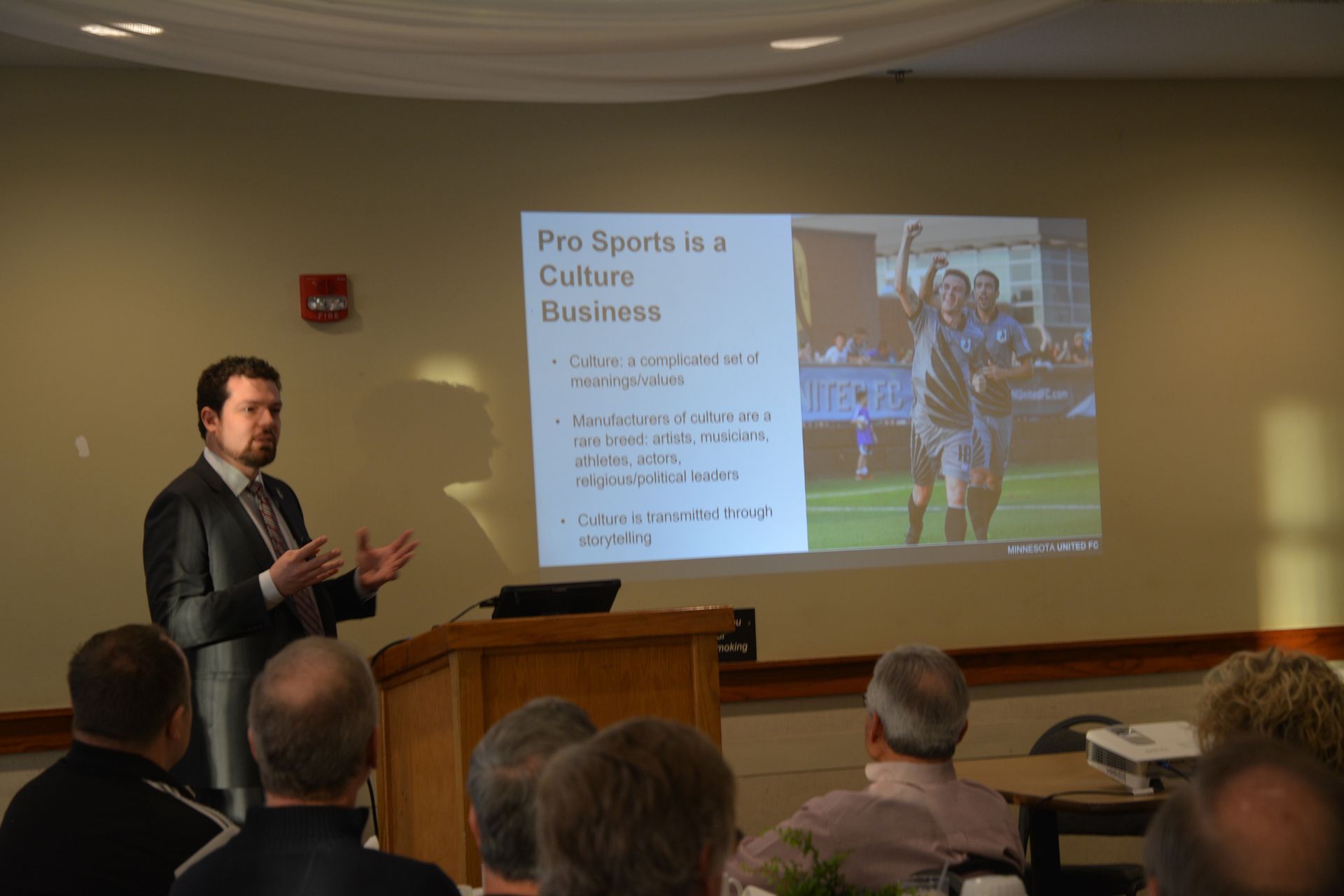 Man giving a presentation, using hand gestures. Slide shows soccer players and text: