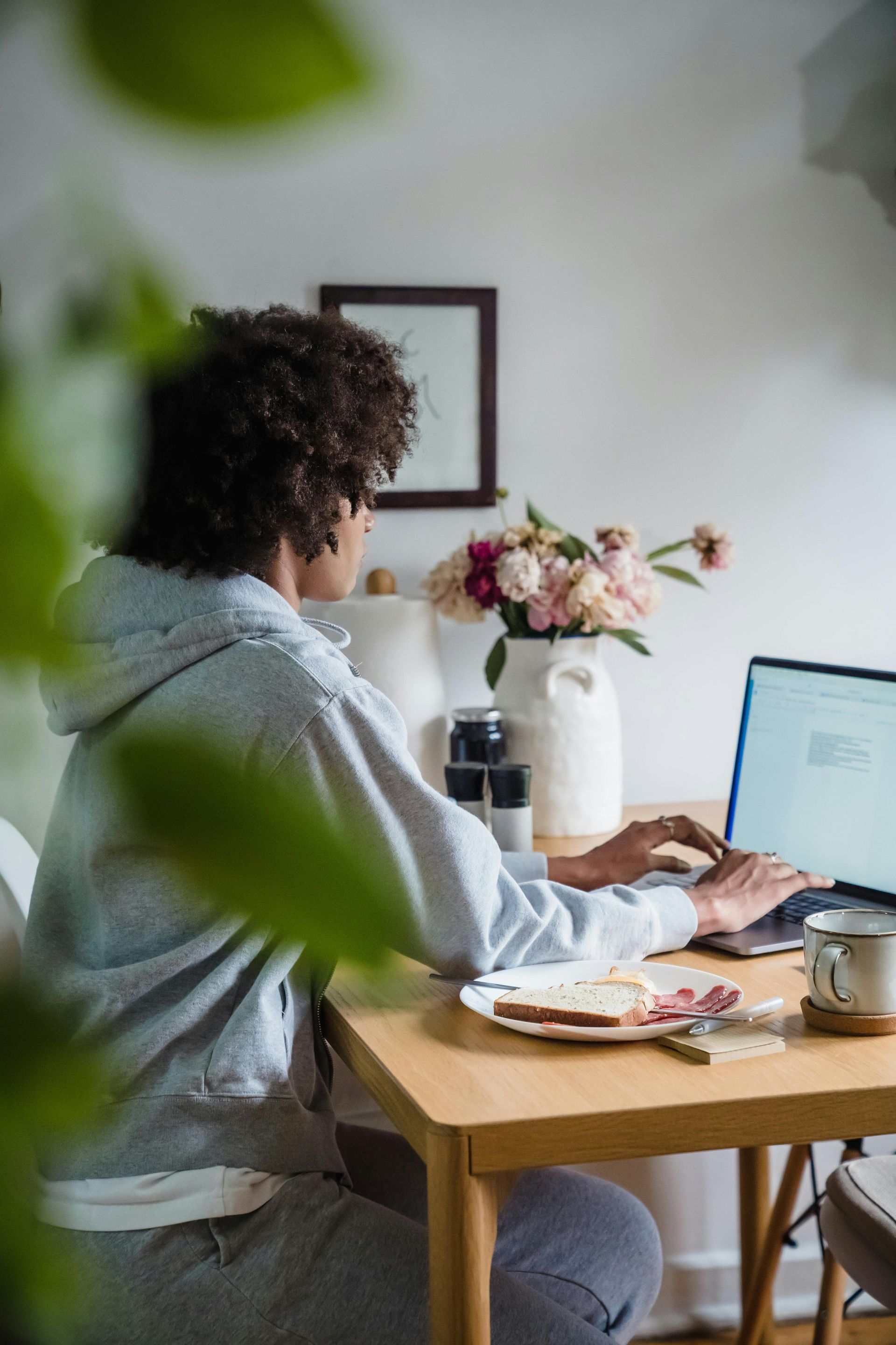 A person in a gray hoodie works on a laptop at a wooden table with a plate of food and a vase of flowers.