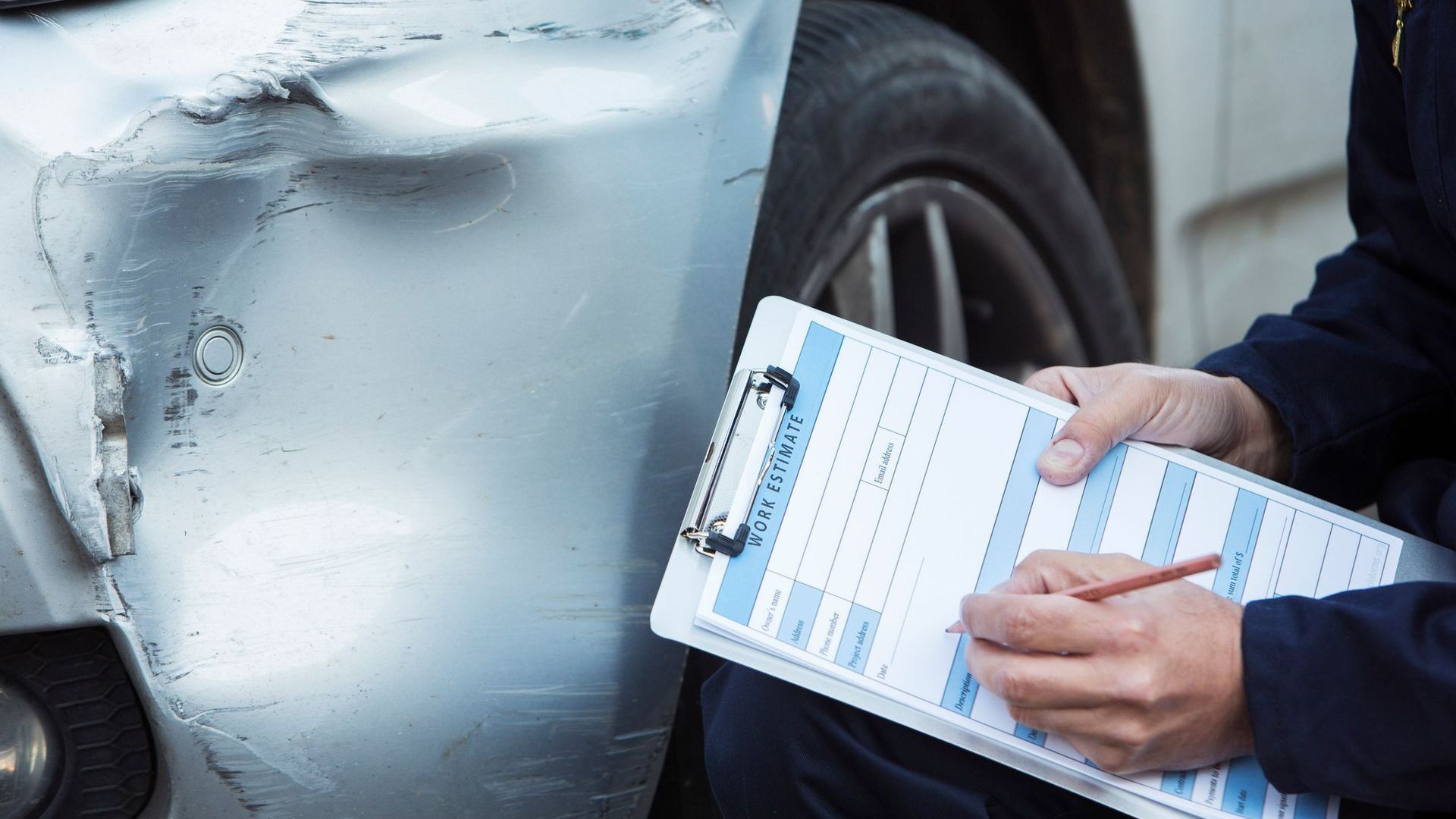 A man is writing on a clipboard in front of a damaged car.