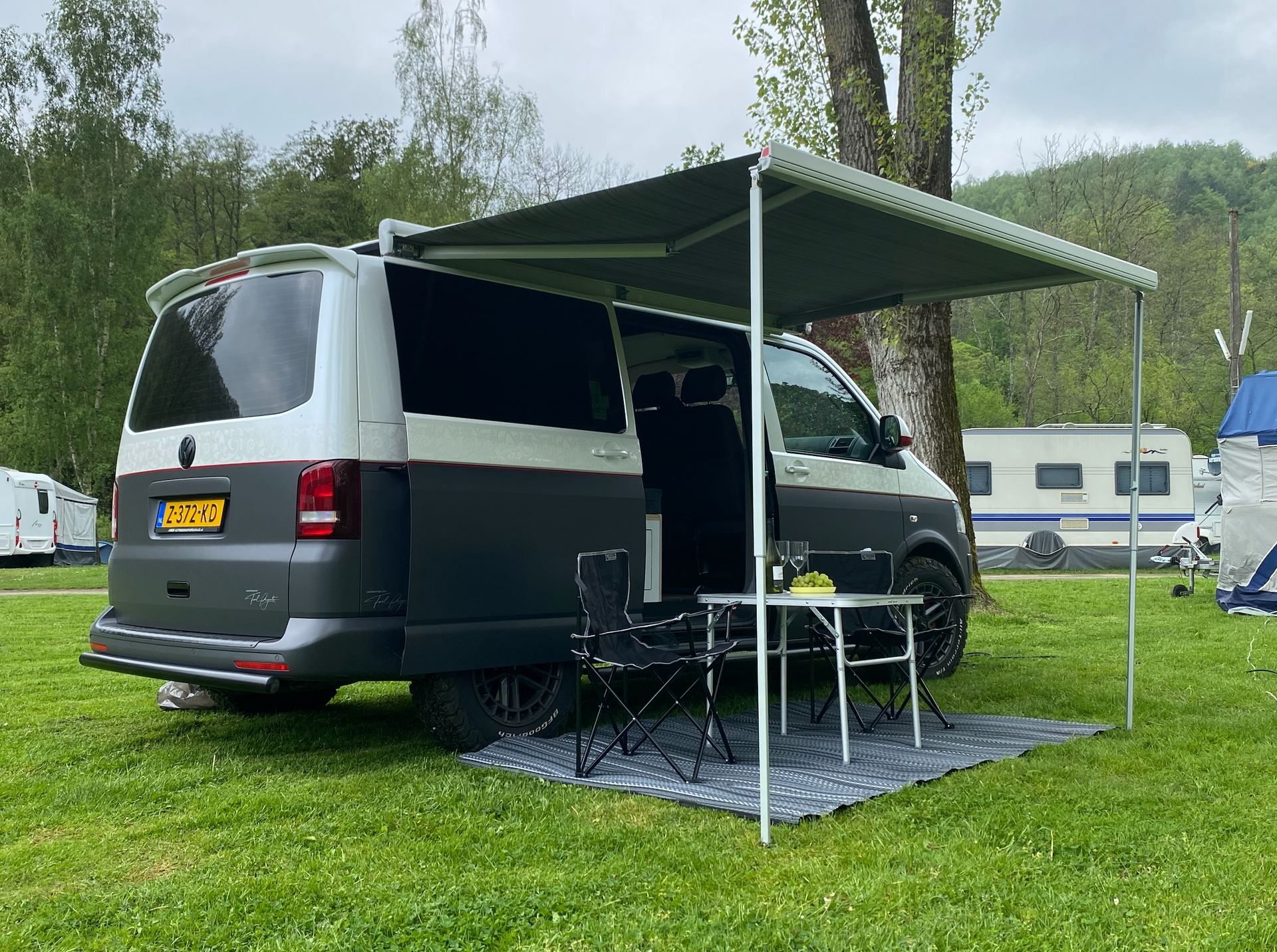 Een busje met luifel staat op een grasveld op de camping; een campingtafel, stoelen en een mat zijn zichtbaar.