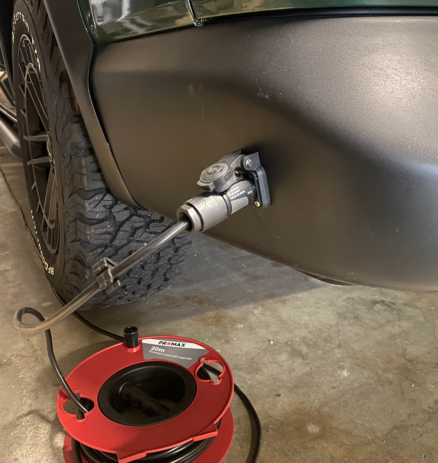 De zijbumper van een zwarte auto met een aangesloten stroomkabel; een rode verlengsnoerhaspel ligt op de grond.