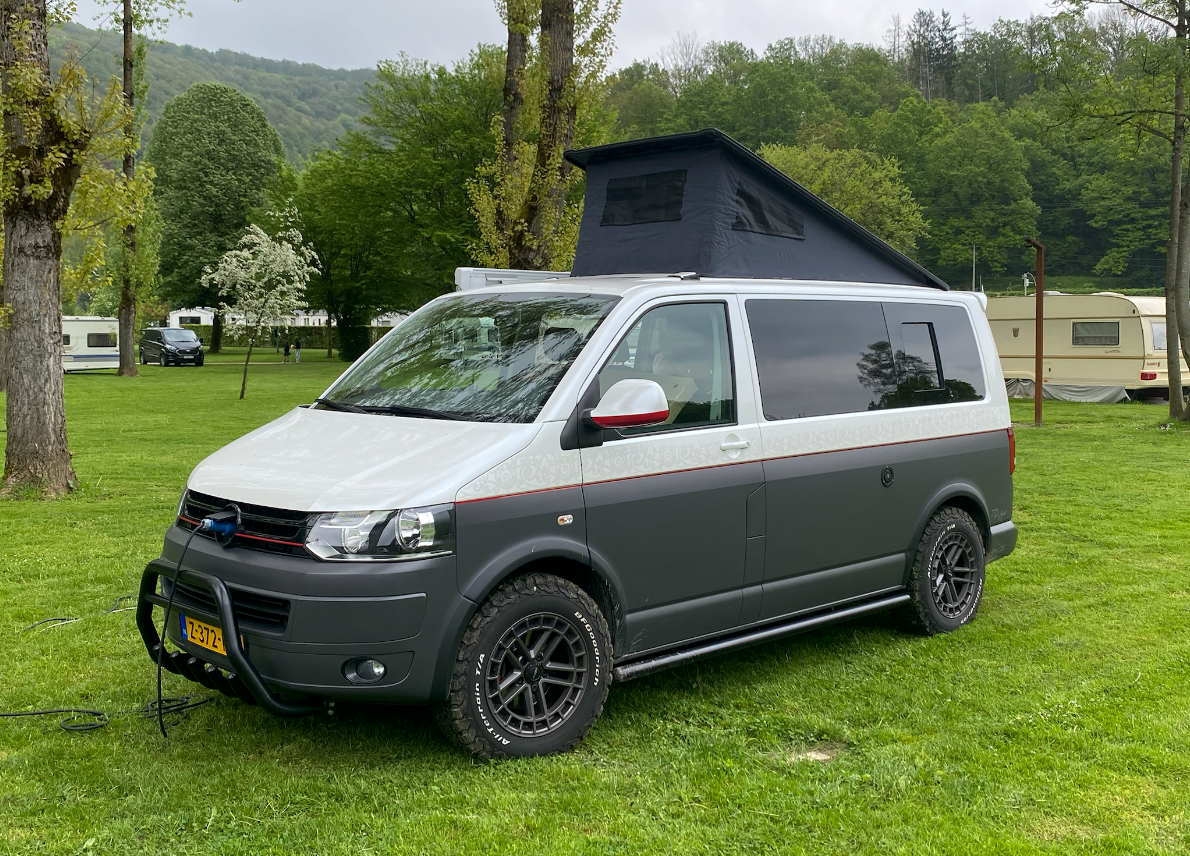 Groene bestelwagen met zonnepanelen op het dak, geparkeerd op een onverharde weg in een natuurlijke omgeving.