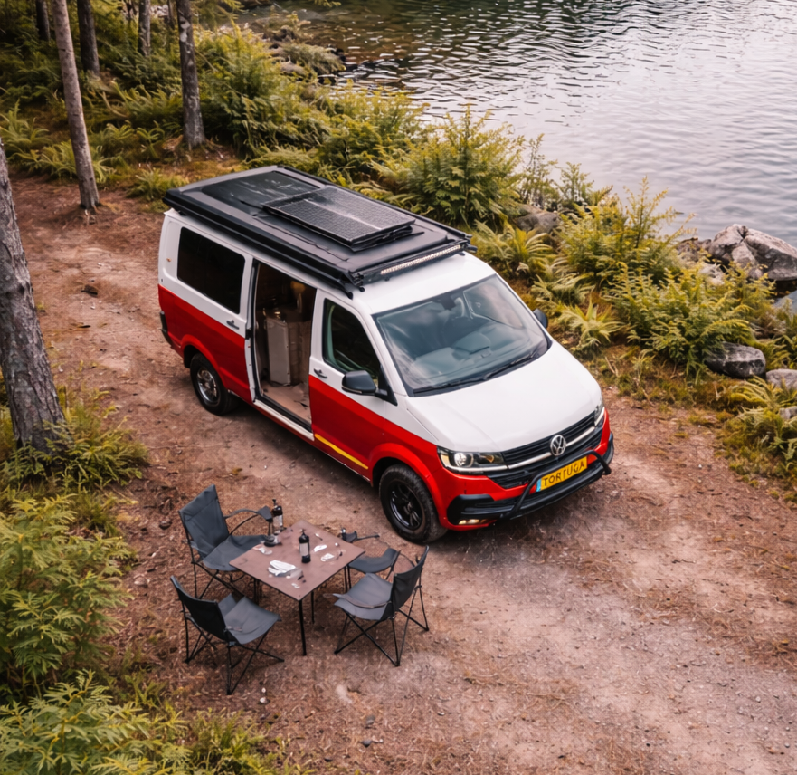 Een rood-witte camper met open deur staat geparkeerd bij een meer, met een tafel en stoelen ernaast.