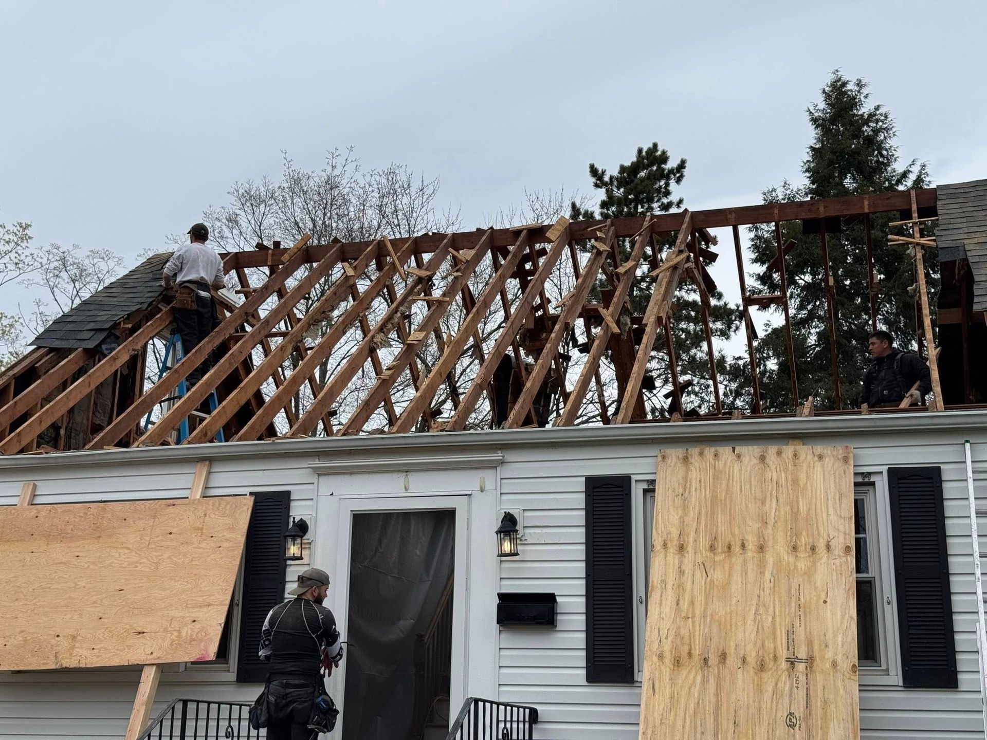 Construction workers replacing a roof on a white house. Rafters exposed, cloudy sky.