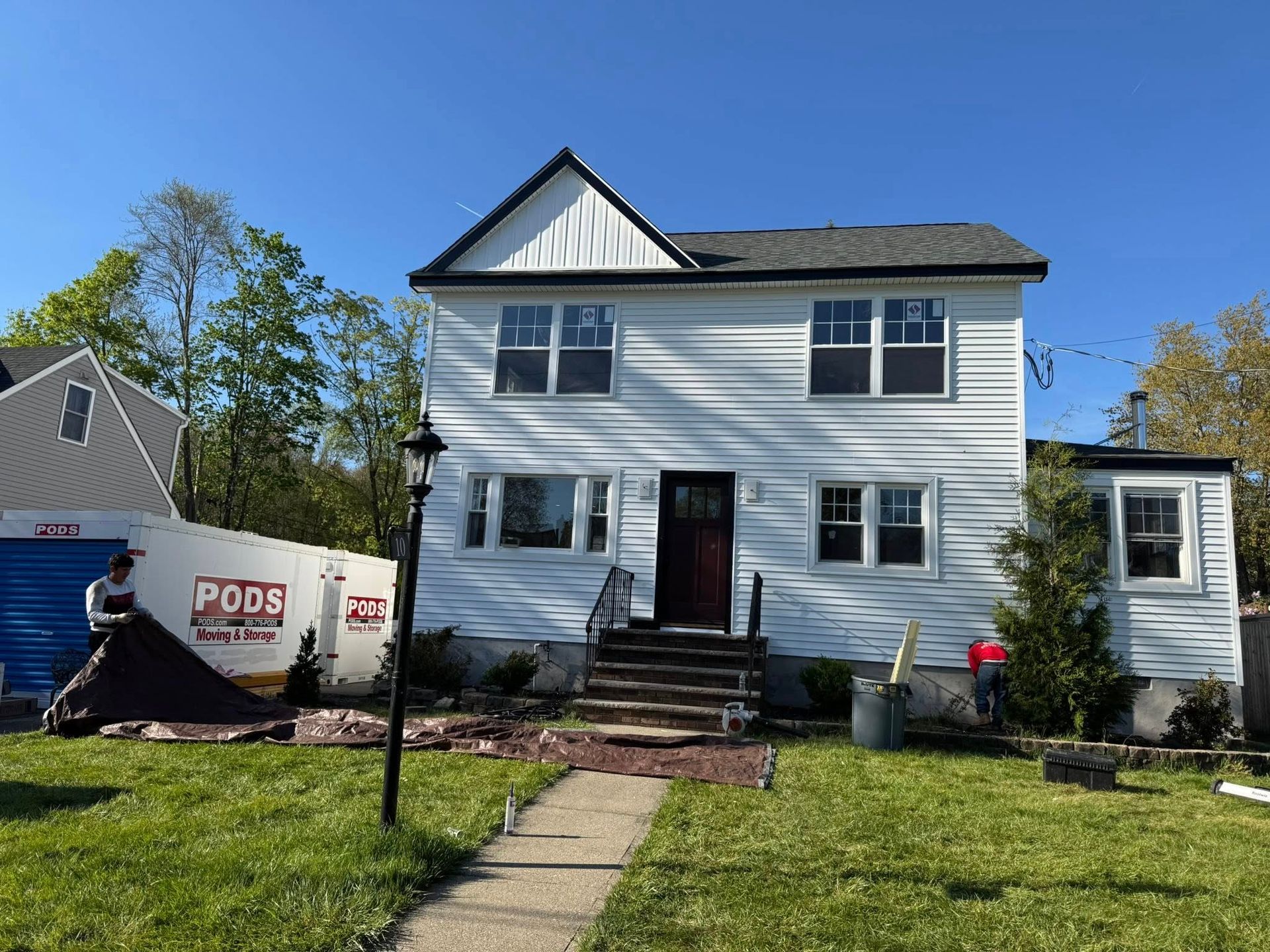White two-story house with black roof, front steps, and a PODS container in the yard on a sunny day.