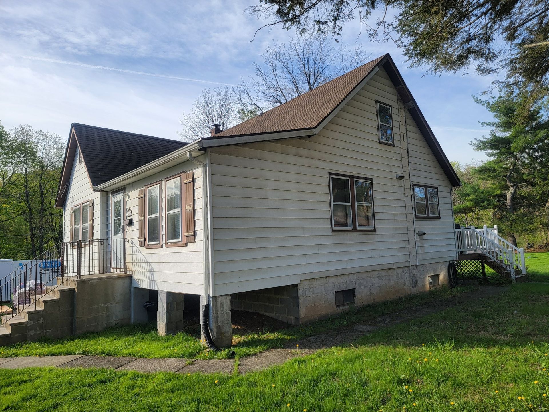 White house with brown roof, shutters, and stairs. Grassy yard, blue sky.