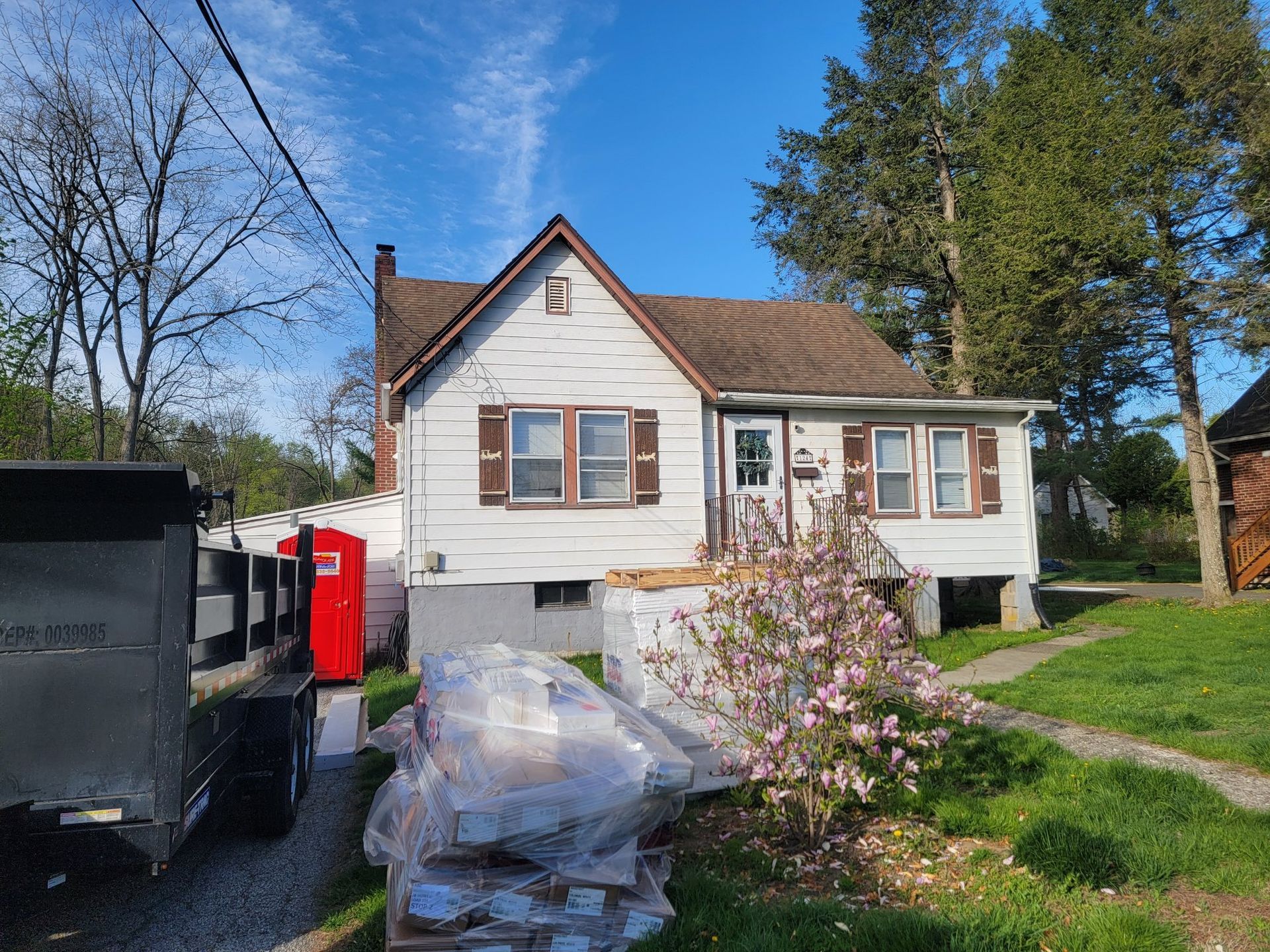 Small white house with brown shutters, a flowering bush, and construction materials.