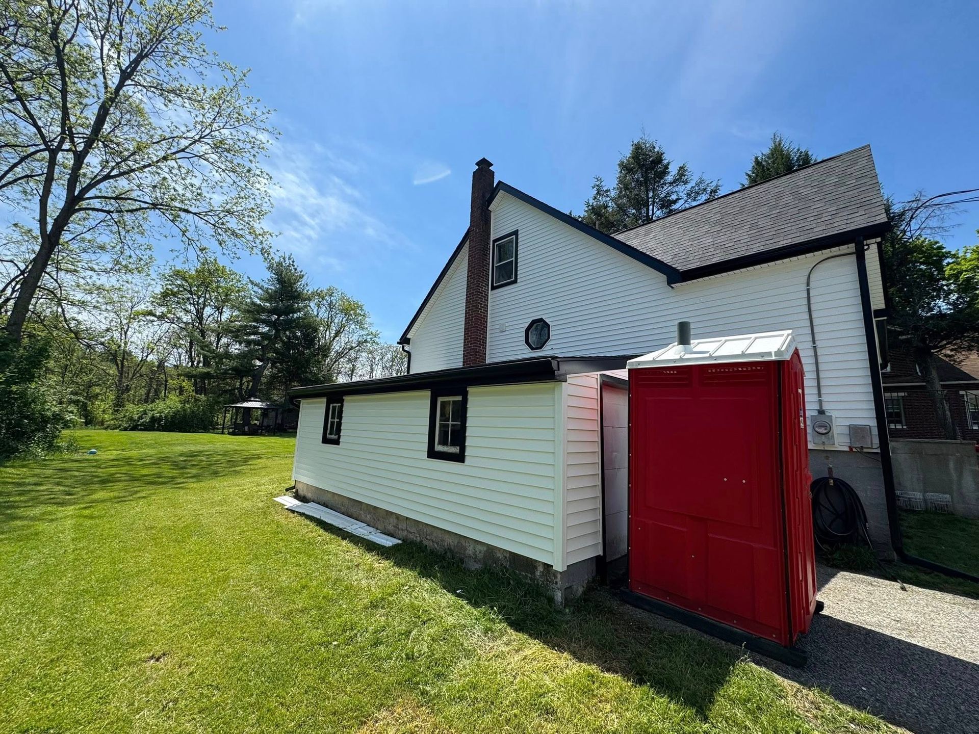 White house with black roof, red portable toilet on side, sunny outdoor setting.