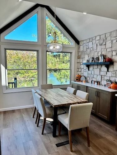 Dining room with a wooden table, six chairs, and large windows overlooking a lake. Stone wall and pumpkins.