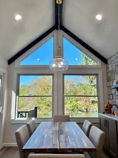 Dining room with large windows, wooden table, and light fixture, overlooking trees and a body of water.