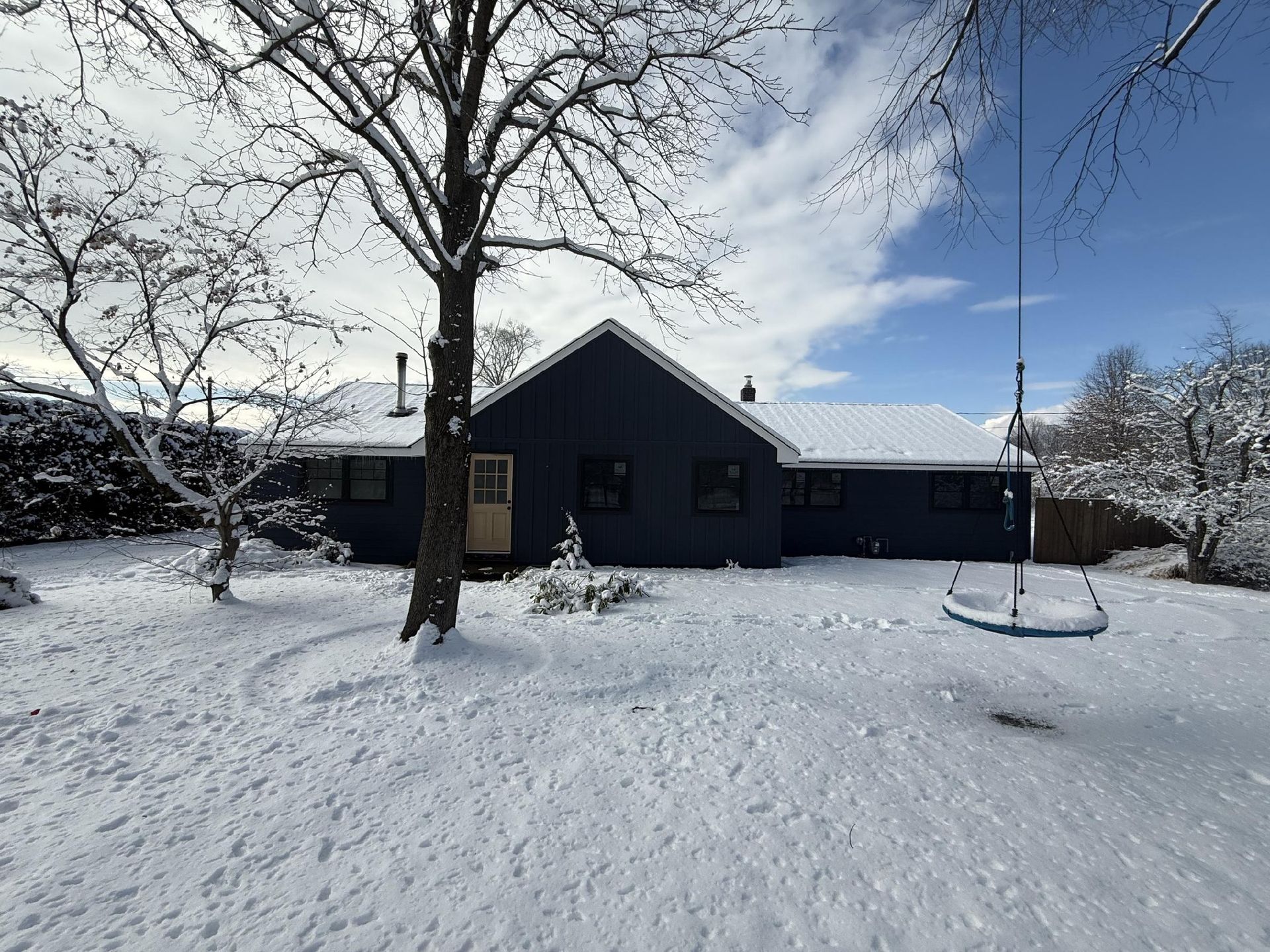 Snow-covered yard with a dark blue house, tree, and swing set under a partly cloudy sky.