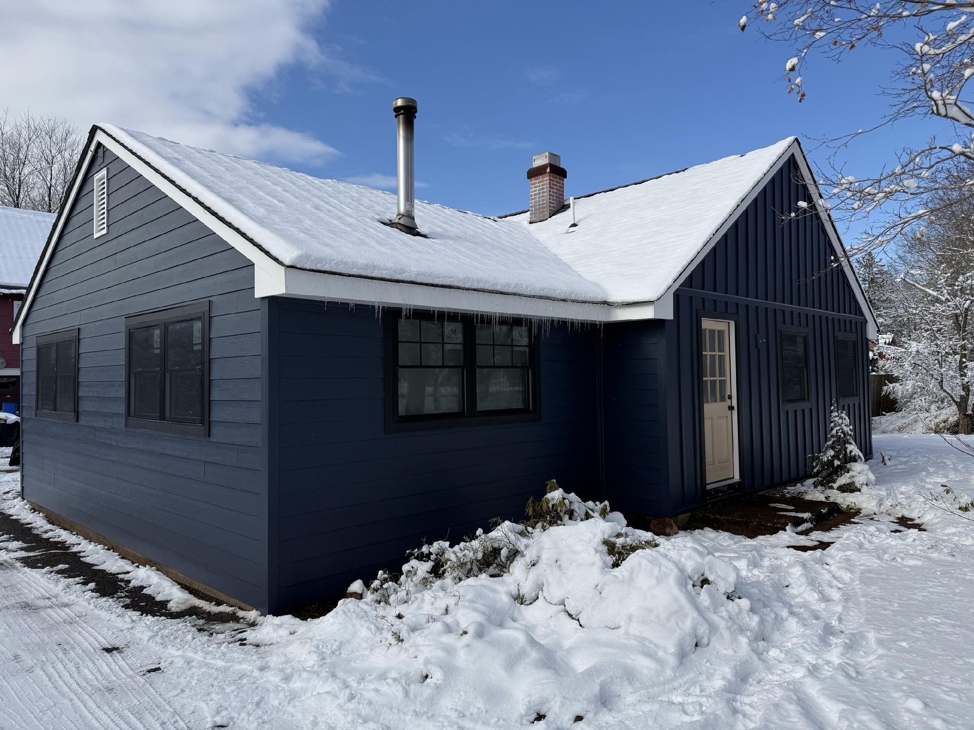 Blue house with snow-covered roof and ground. Chimneys, windows, and a doorway are visible. Sunny day.
