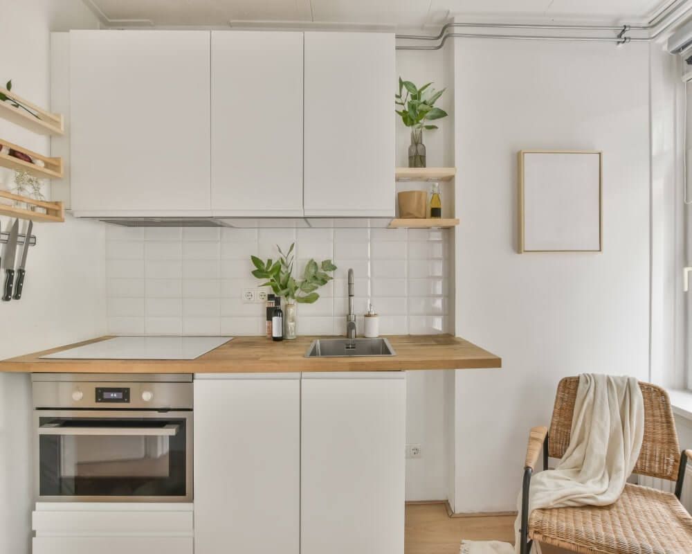 Small, white kitchen with wooden countertop, built-in oven, sink, and decorative plants.