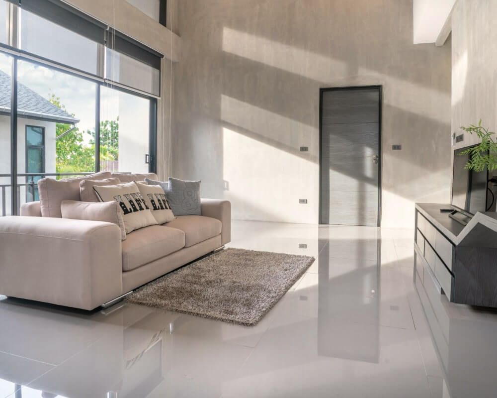 Living room with beige sofa, rug, and large windows; bright sunlight reflects on glossy floors.