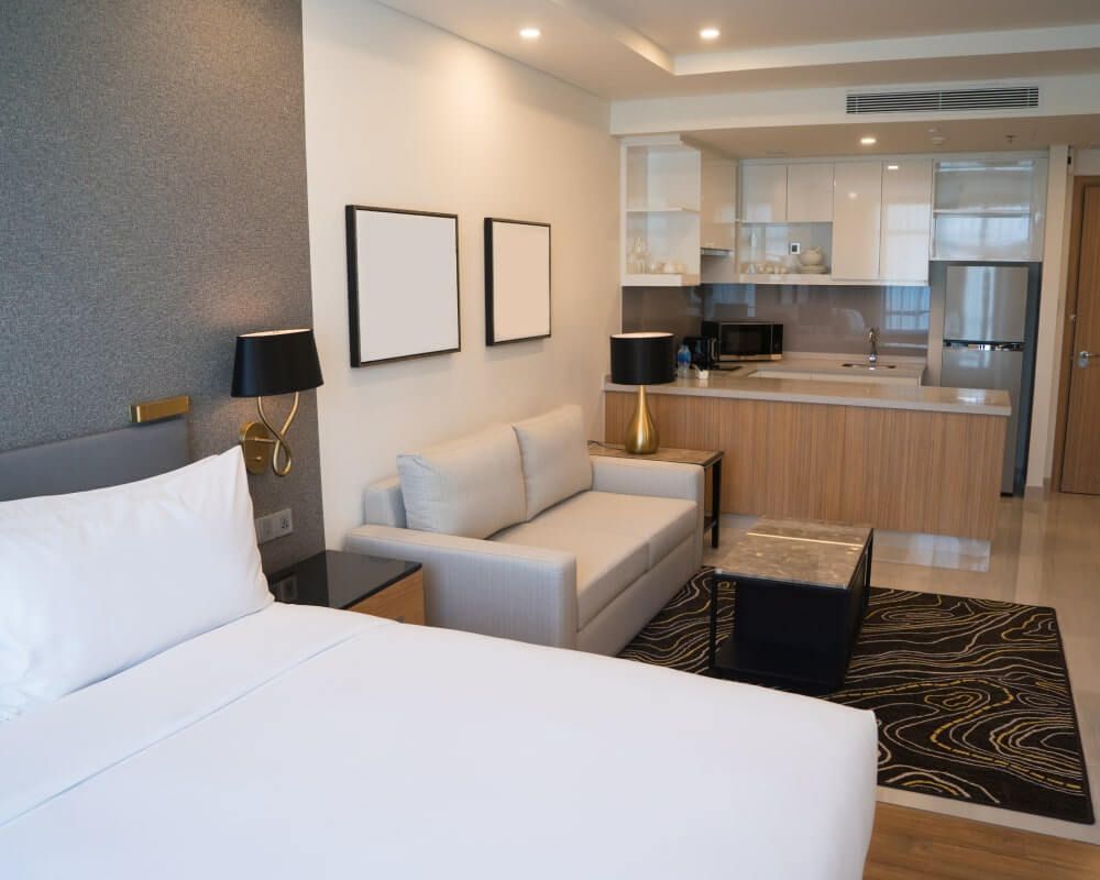 Modern hotel room with a bed, small sofa, kitchenette, and artwork.