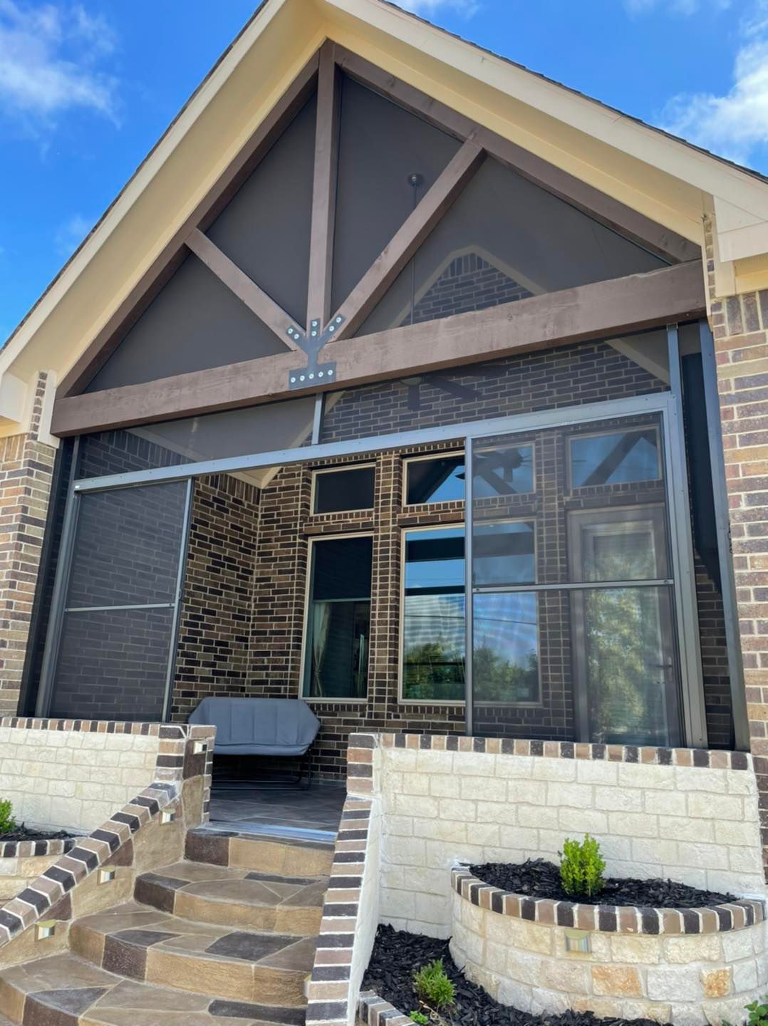 A screened-in patio with a gabled roof, stone facade, and steps leading to a seating area under a bright blue sky.