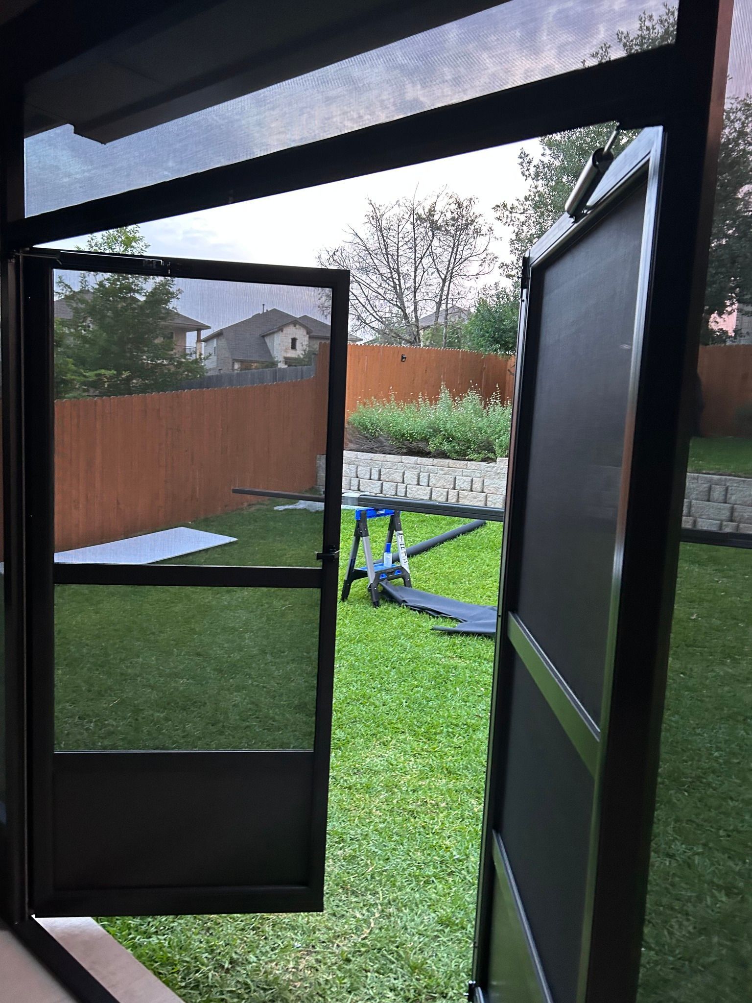 A view through a set of open black screen doors, revealing a green backyard with a fence and a small toy set on the lawn.