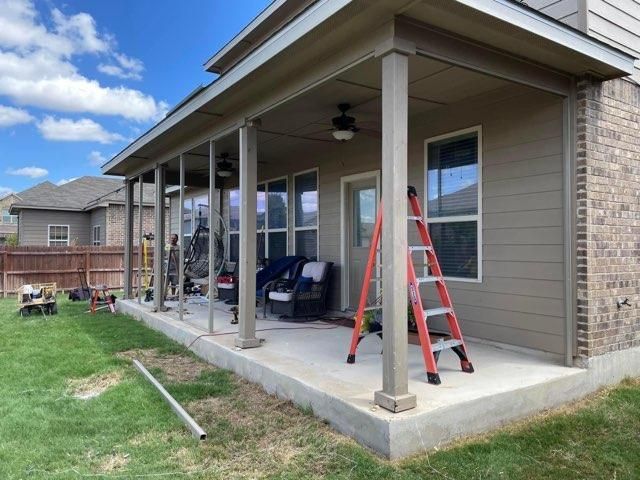A backyard view showing a concrete patio with a covered porch, outdoor furniture, two ceiling fans, and an orange ladder.
