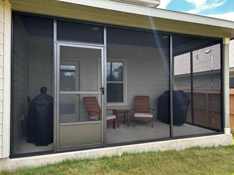 A screened-in porch with a door, two wooden chairs, and two covered grills on a concrete patio beside a house.